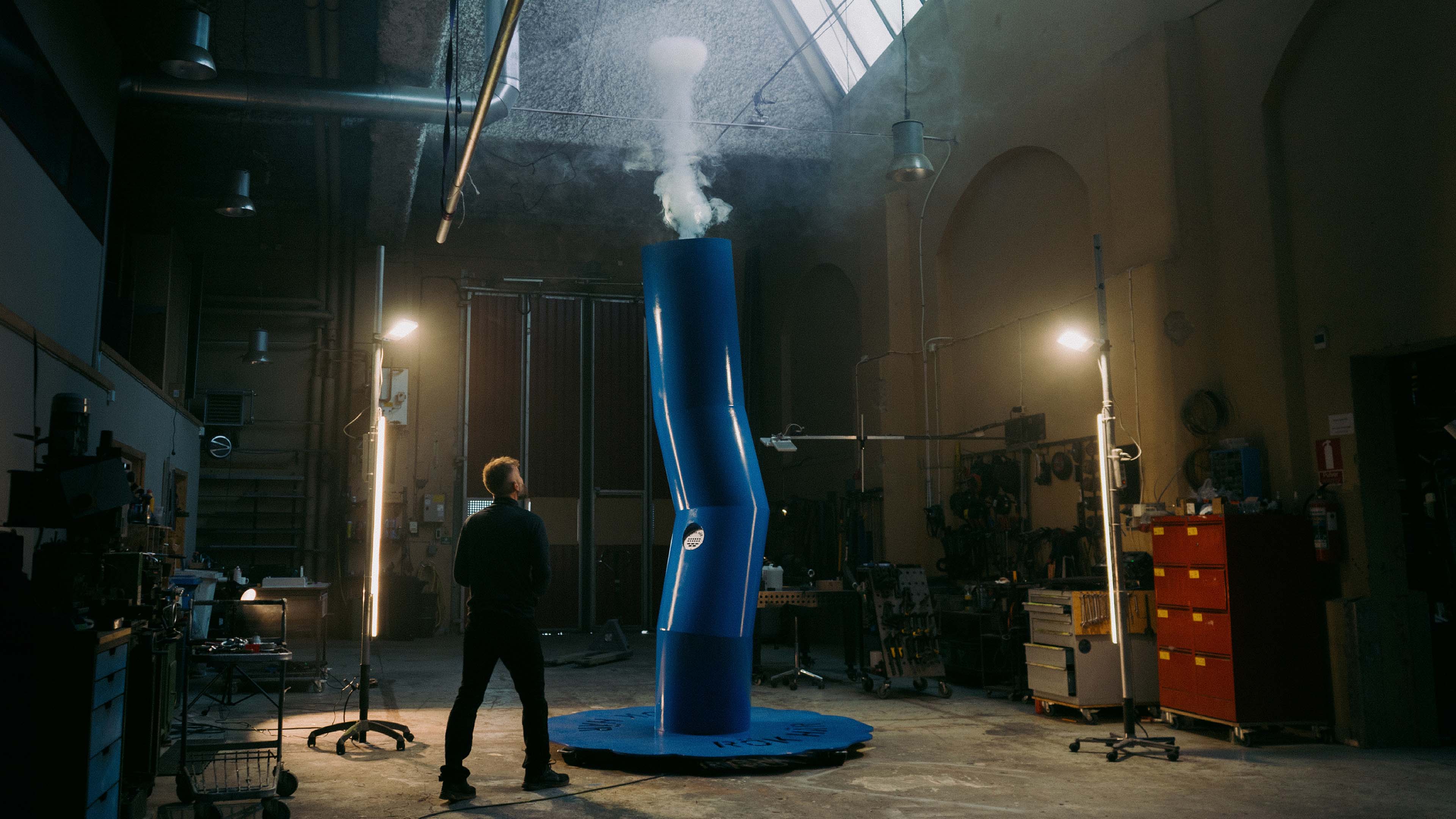 Person standing in a large industrial workshop next to a tall blue cylindrical machine emitting white smoke upwards.