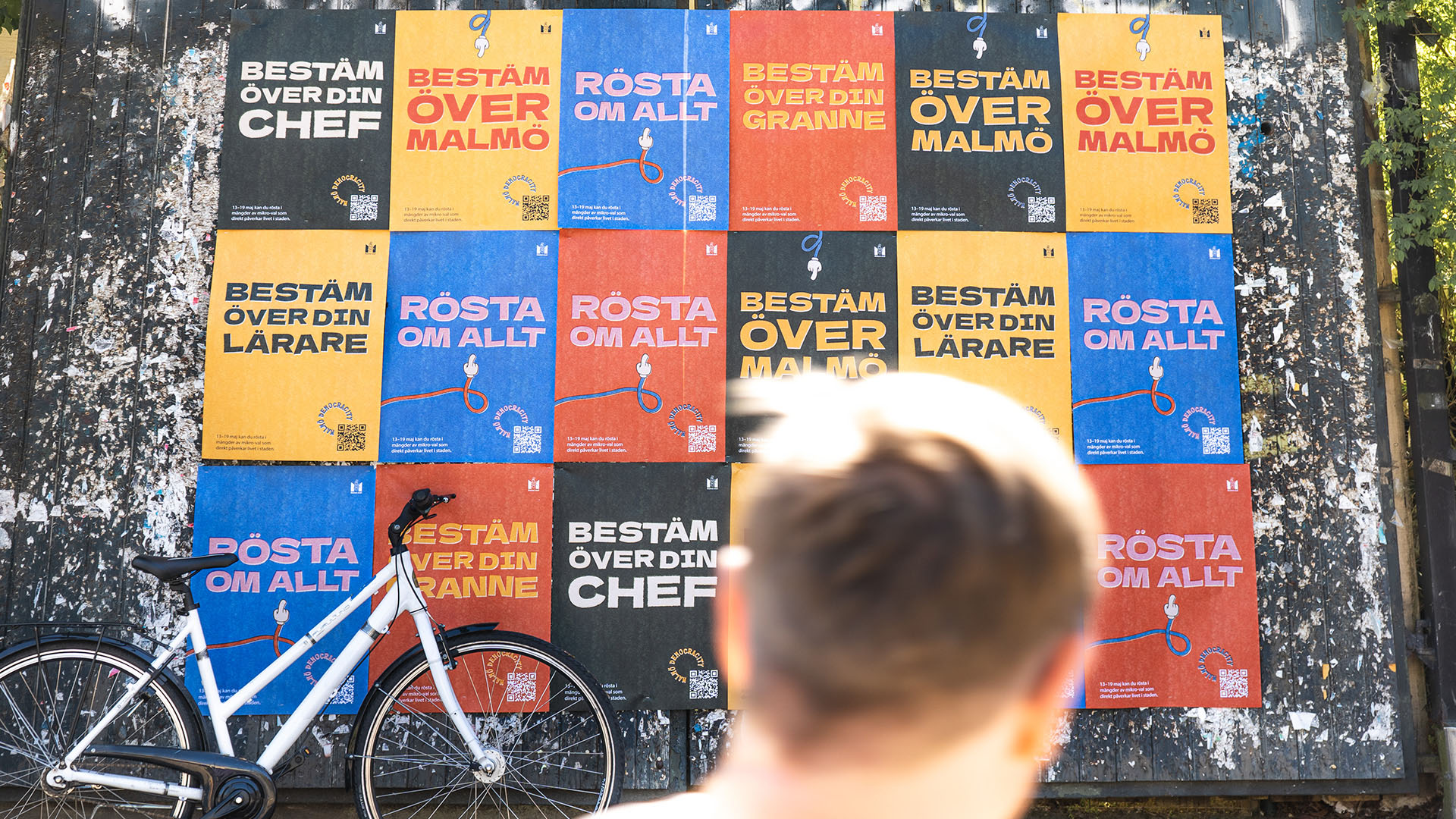 Wall covered with colorful posters in Swedish urging voting on various local issues, with a bicycle in front and a person looking at the wall.