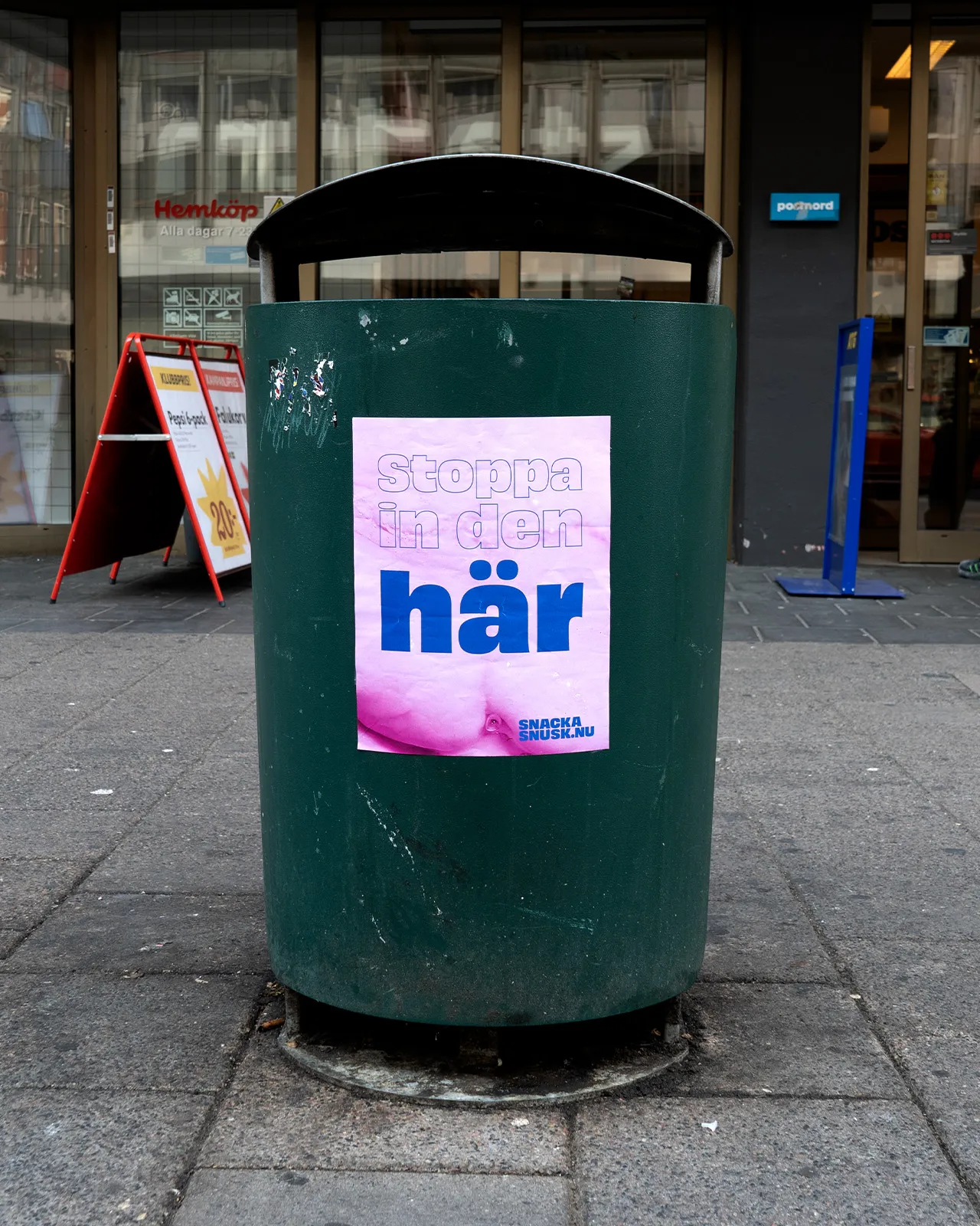 Green trash bin on a city sidewalk with a pink poster displaying Swedish text 'Stoppa in den här'.