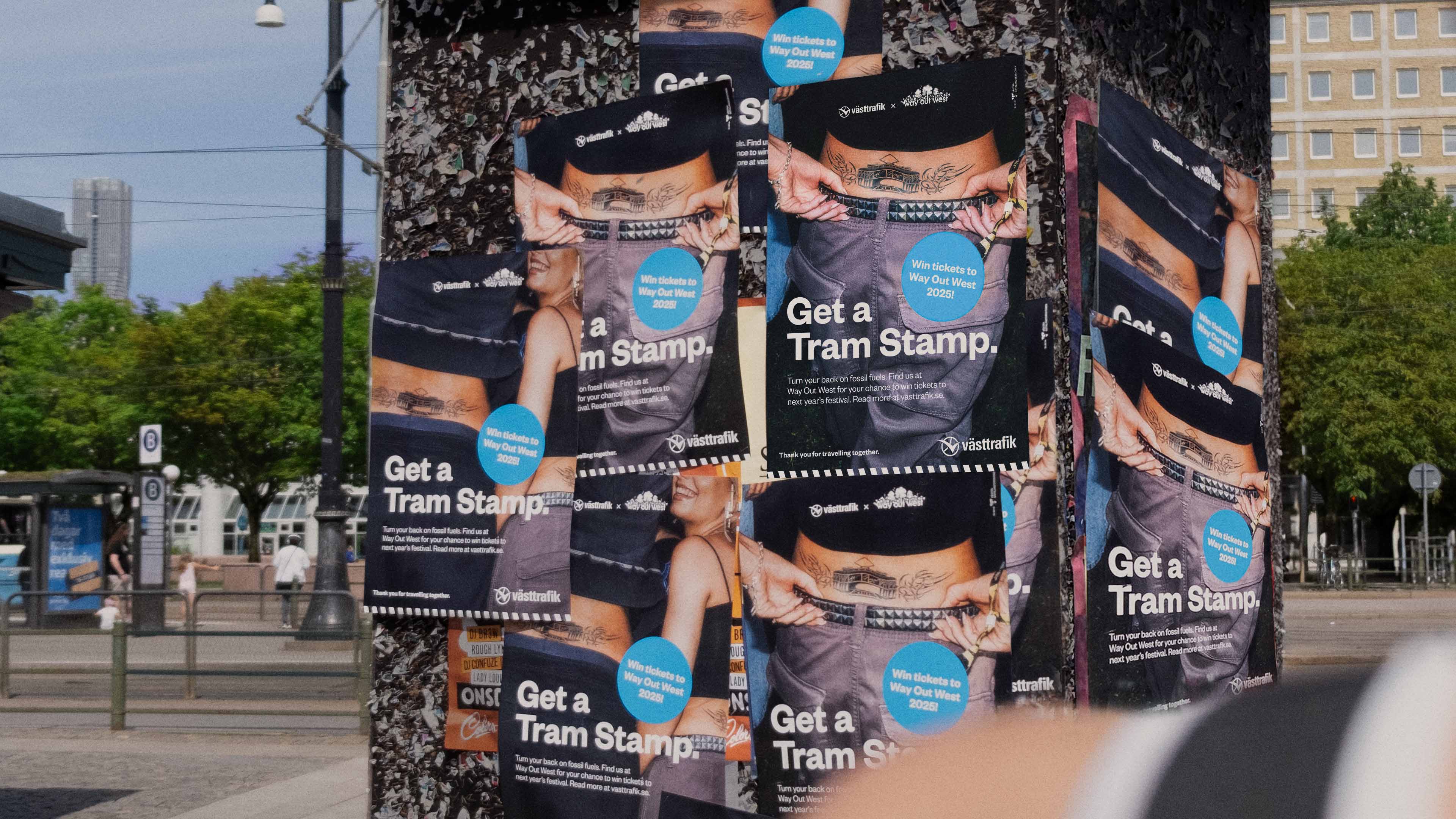 Street pole covered with repeated posters advertising 'Get a Tram Stamp' offering tickets to Way Out West 2025 with image of a person showing tattoo and studded belt.