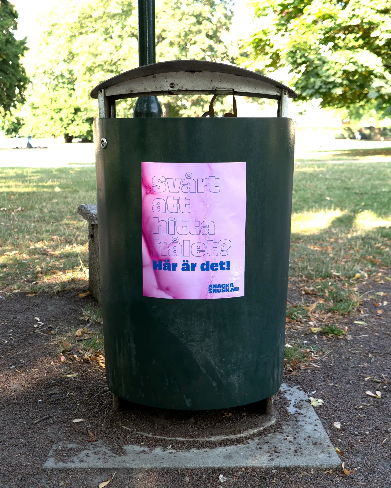 Green outdoor trash bin with a pink poster that reads 'Svårt att hitta hålet? Här är det!' attached to the front.