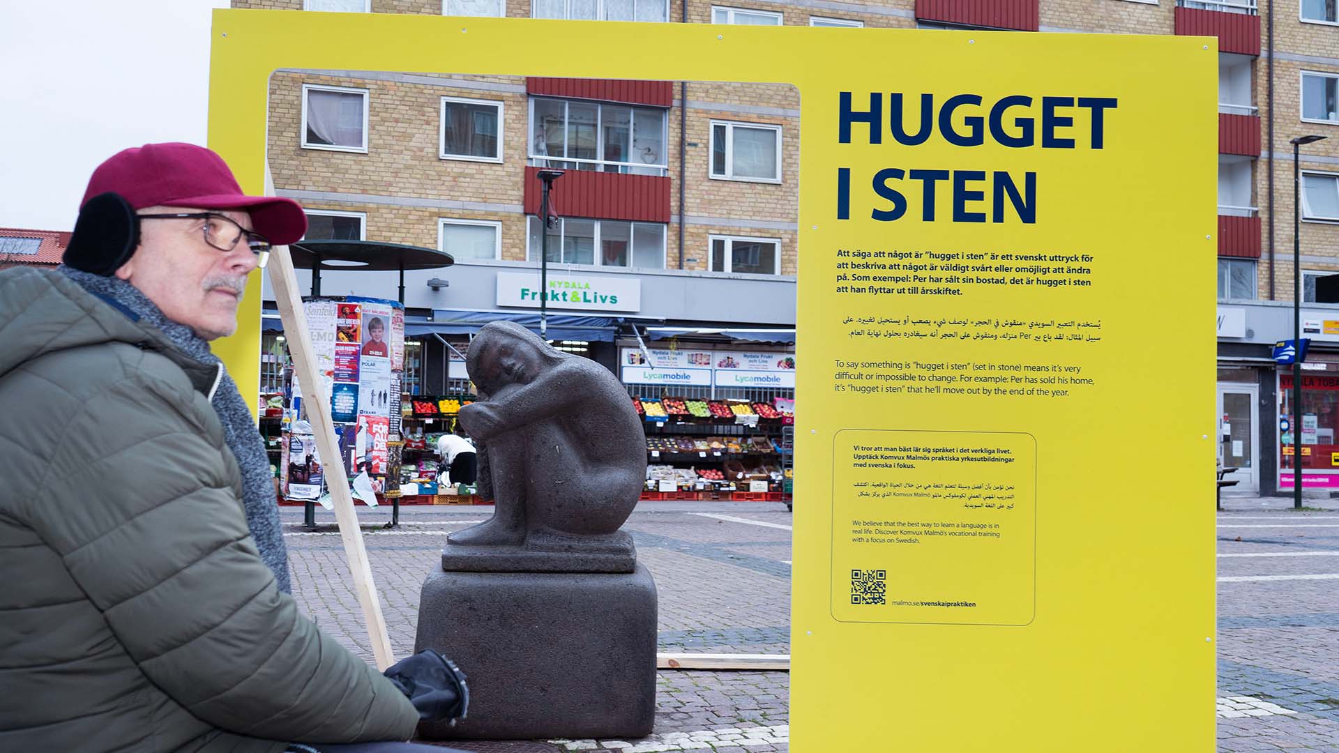 Man in warm clothing sitting near a stone sculpture of a curled-up figure, with a large yellow sign explaining the Swedish phrase 'Hugget i sten' in the background.
