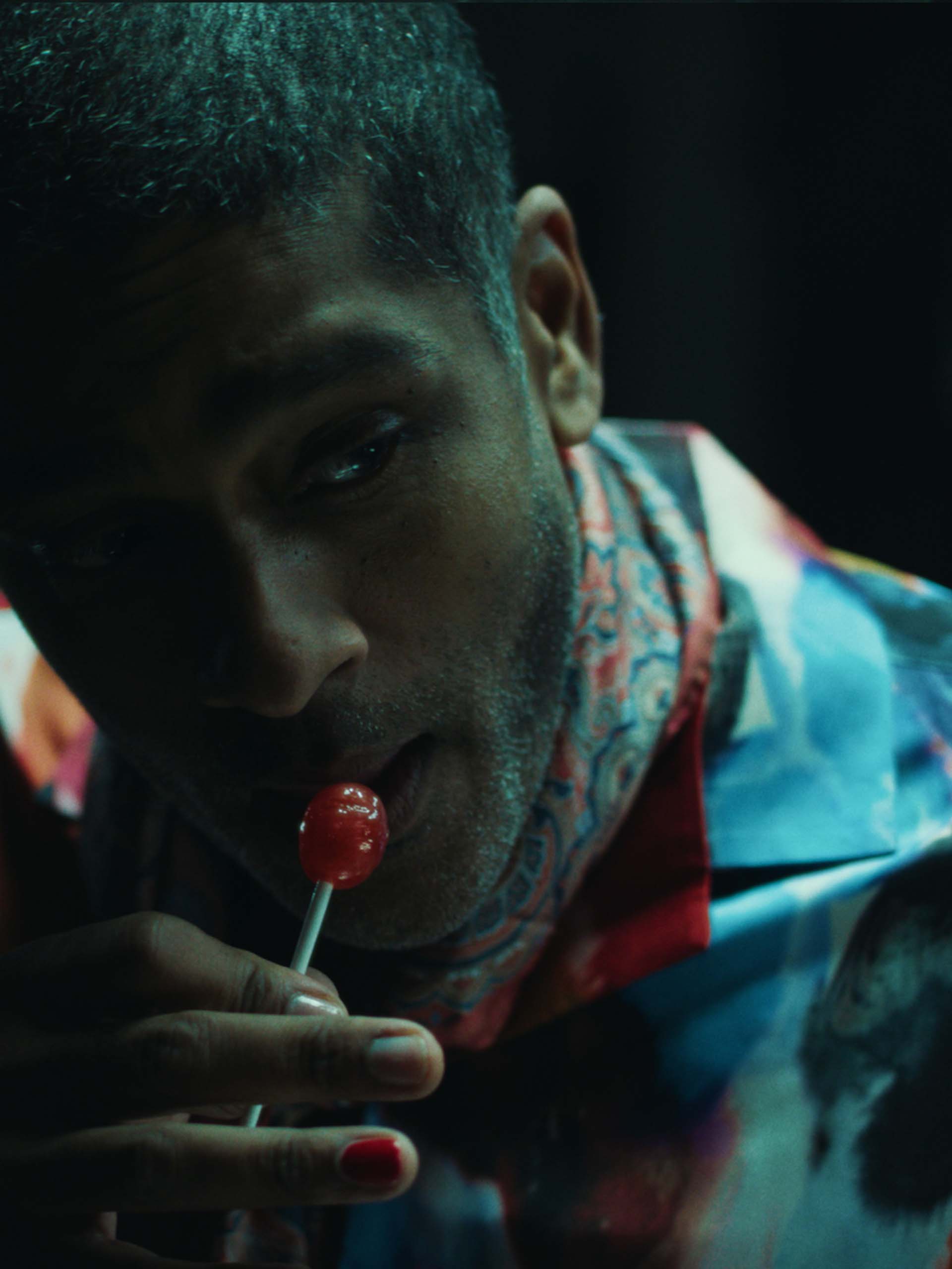 Man in colorful shirt and scarf holding a red lollipop near his mouth in dim lighting.