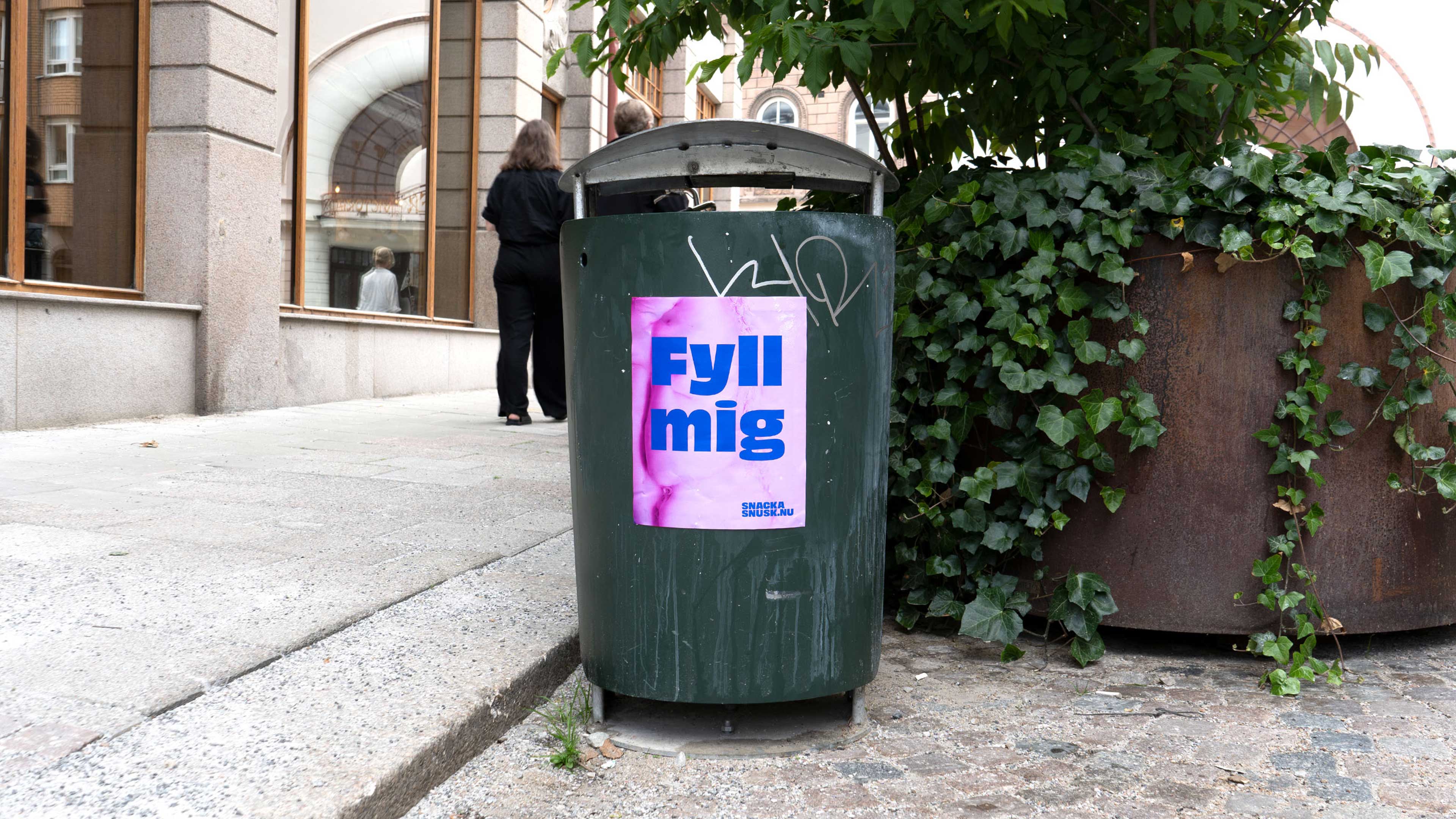 Green outdoor trash bin with a pink sign reading 'Fyll mig' and ivy-covered planter beside it.