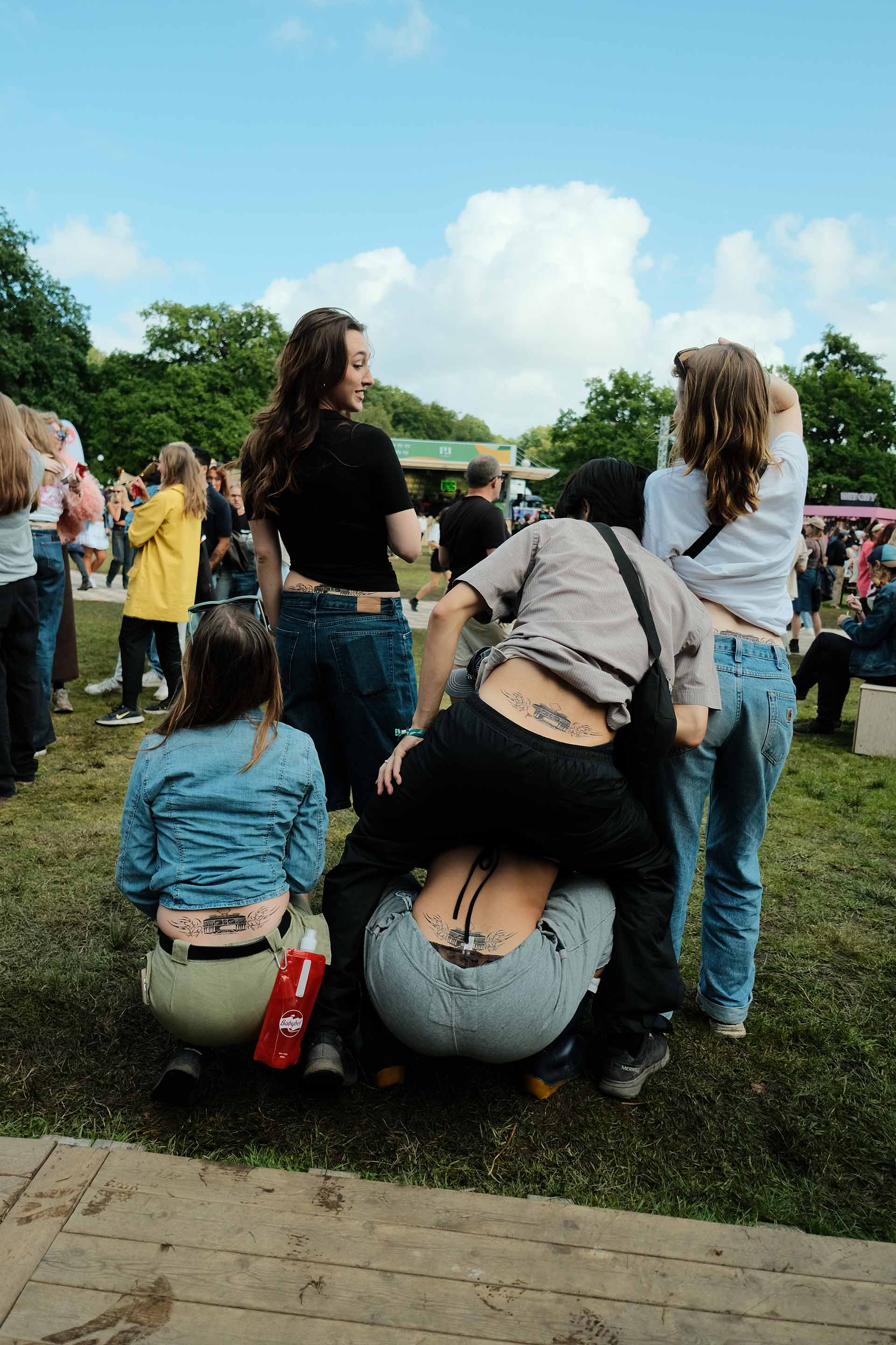 Group of five young people at an outdoor event showing matching lower back tattoos while squatting or standing on grass with trees and crowd in background.
