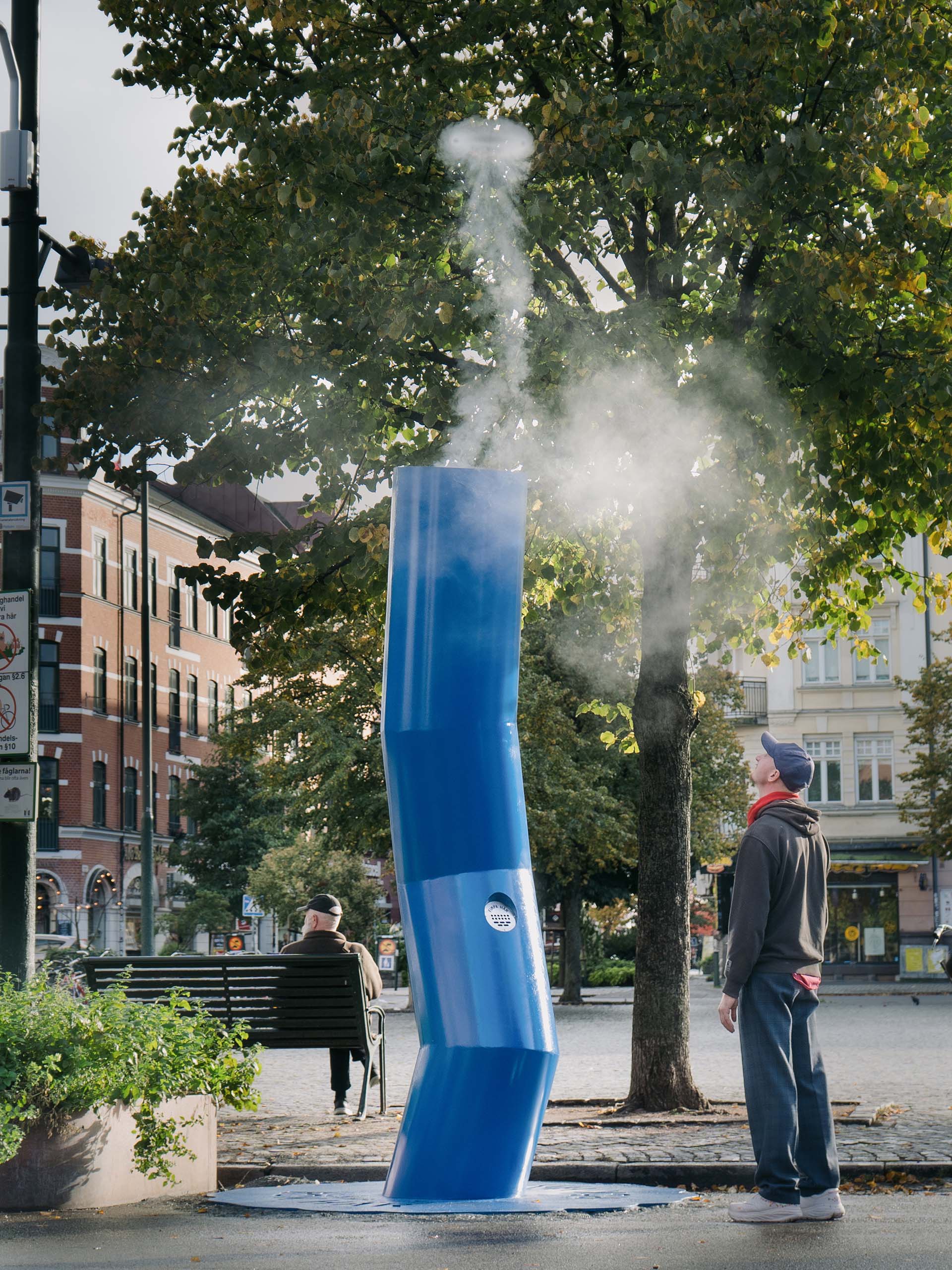 A person stands outdoors looking up at a large blue sculpture shaped like a bent cigarette emitting smoke, with a park bench and trees in the background.