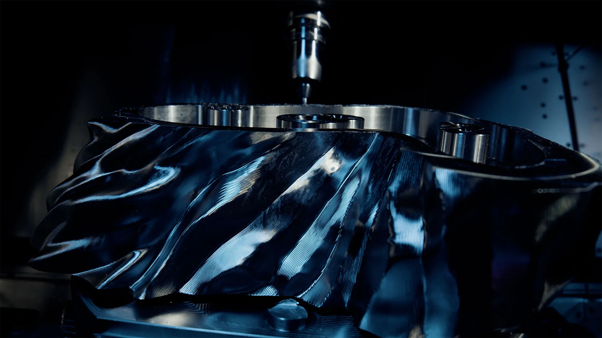 Close-up of a shiny, intricately machined metal component under a precision milling machine tool in a dark workshop.