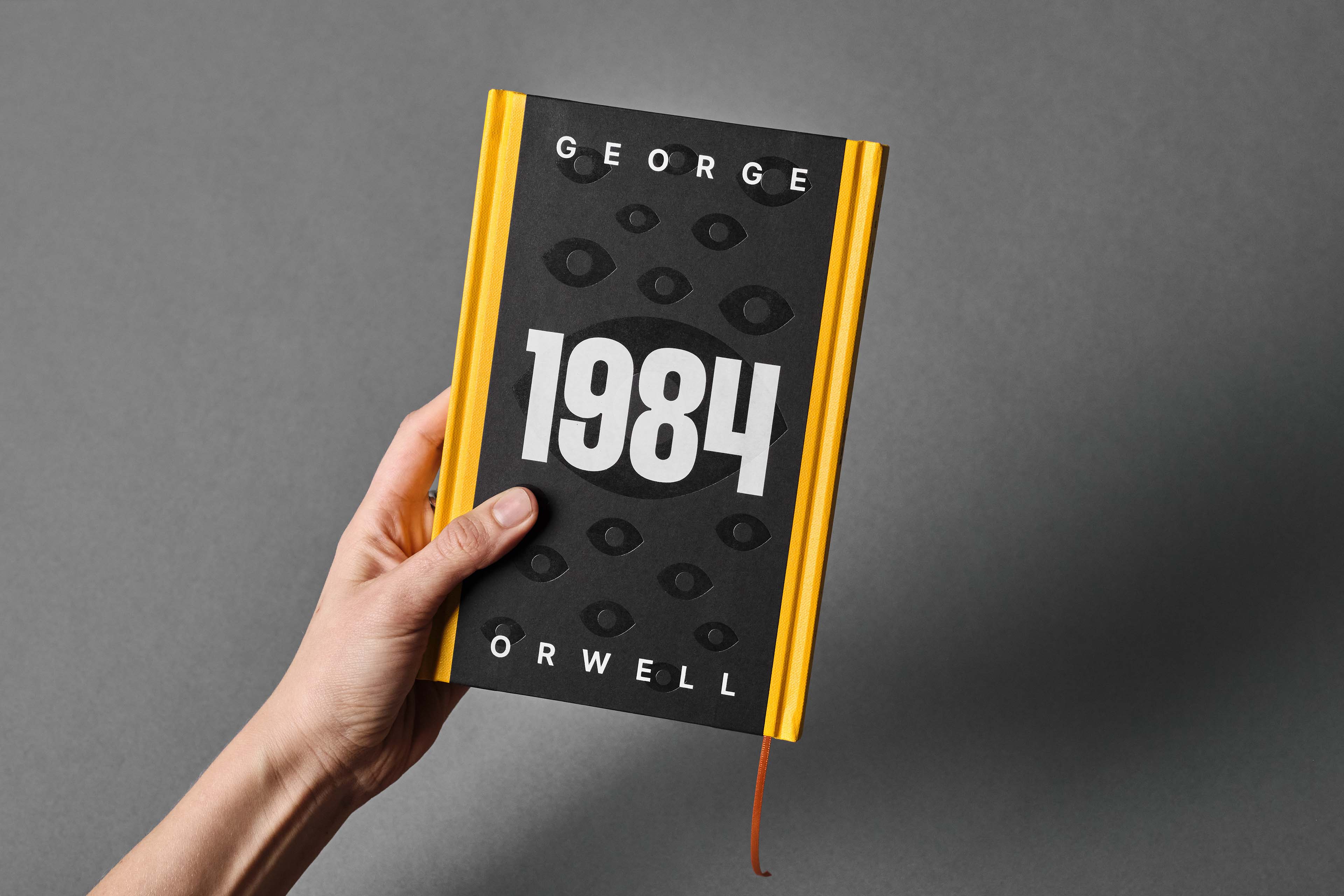 Hand holding a hardcover edition of George Orwell's book 1984 with a black and yellow cover featuring multiple eye illustrations.