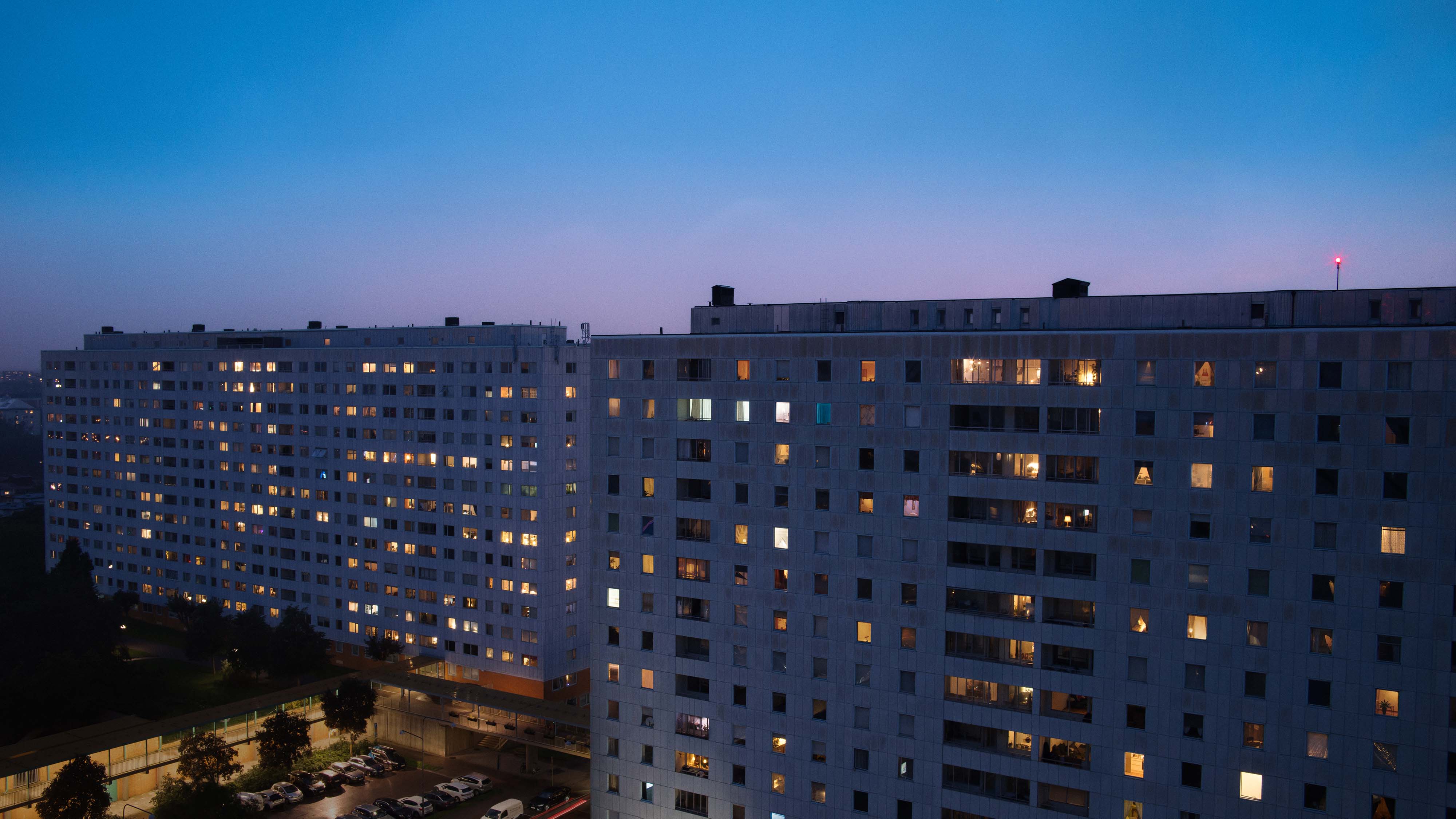 Two large apartment buildings lit from many windows at dusk under a clear sky.