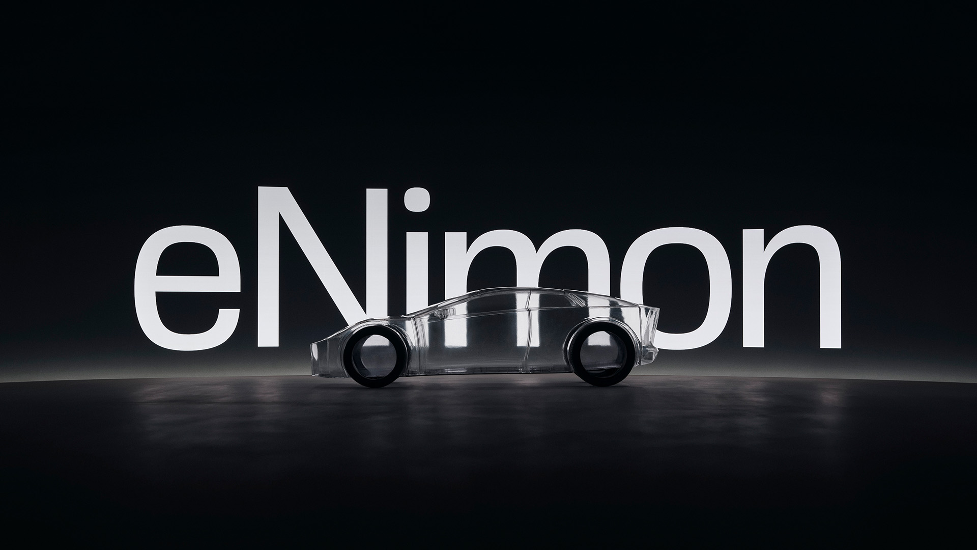 Transparent model of a car with black wheels placed in front of large illuminated text reading 'eNimon' on a dark background.