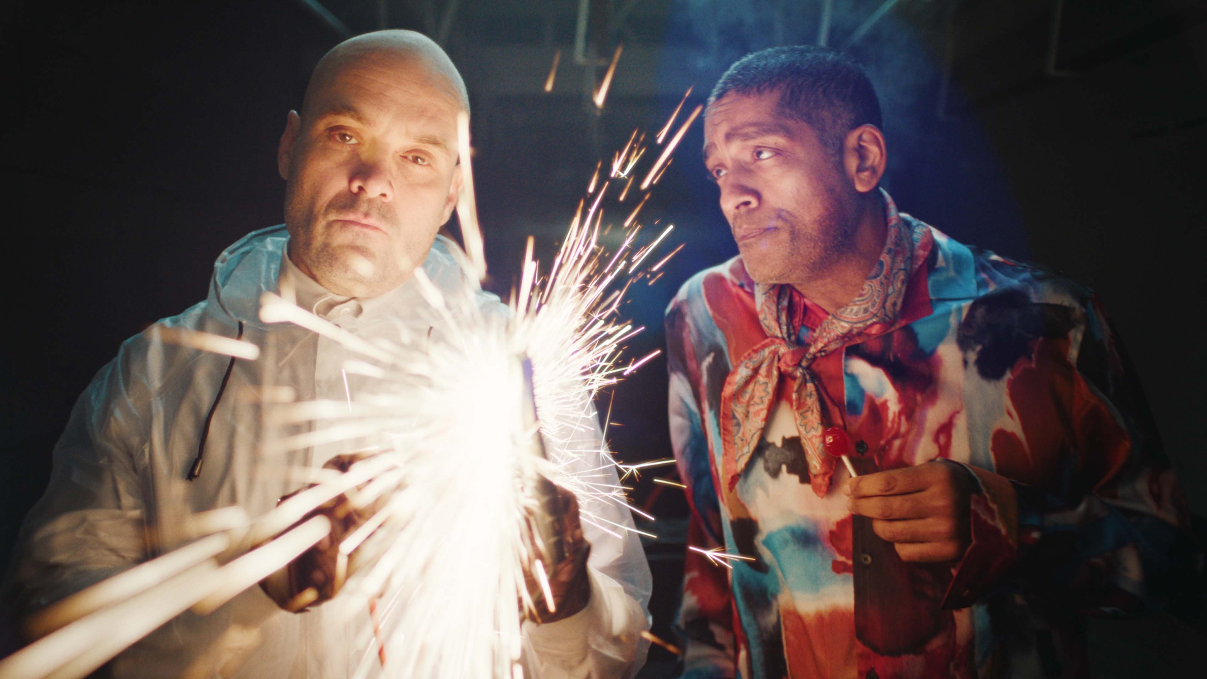 Two men in colorful clothing holding sparklers with sparks flying in a dark setting.