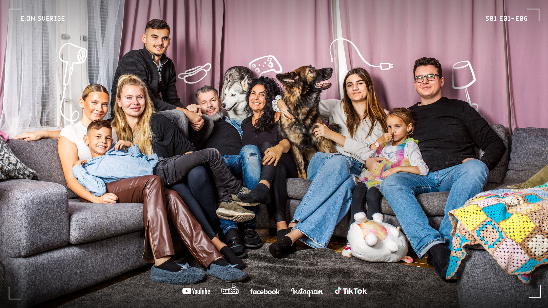 A multigenerational family of eight and two large dogs sitting closely together on a gray sectional sofa in a cozy living room with purple curtains.