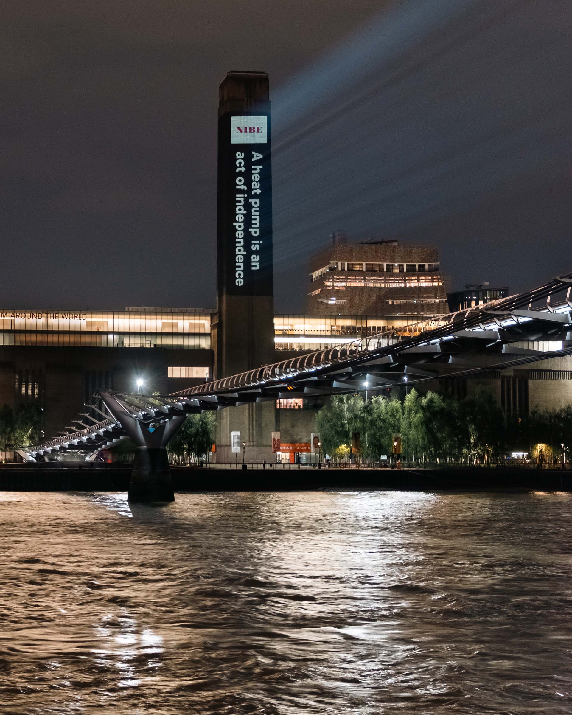 Night view of a lit bridge over water with a tall building displaying a heat pump message in the background.