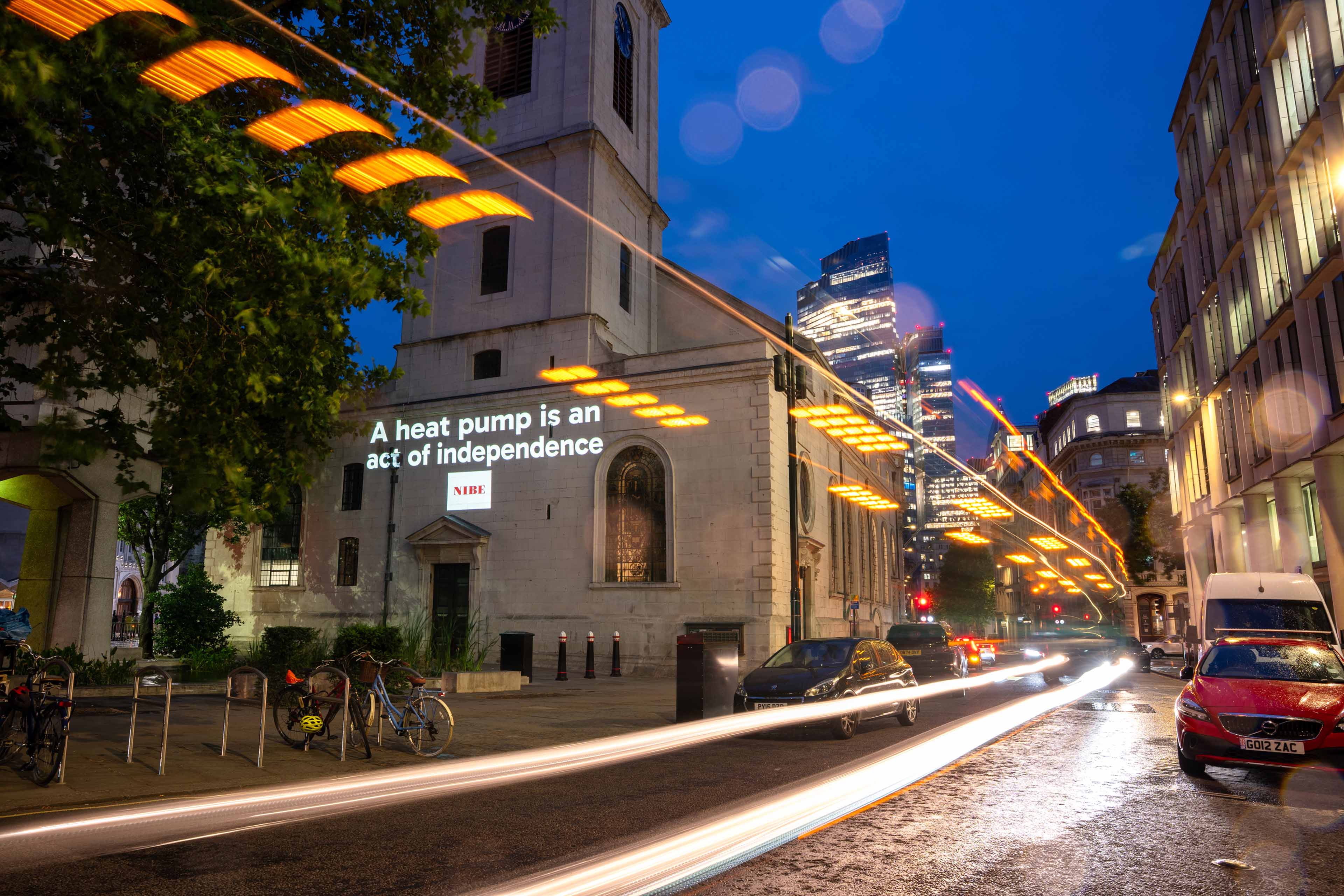 City street at night with light trails, buildings, parked cars, and a church with illuminated text on its wall "A heat pump is and act of independence"