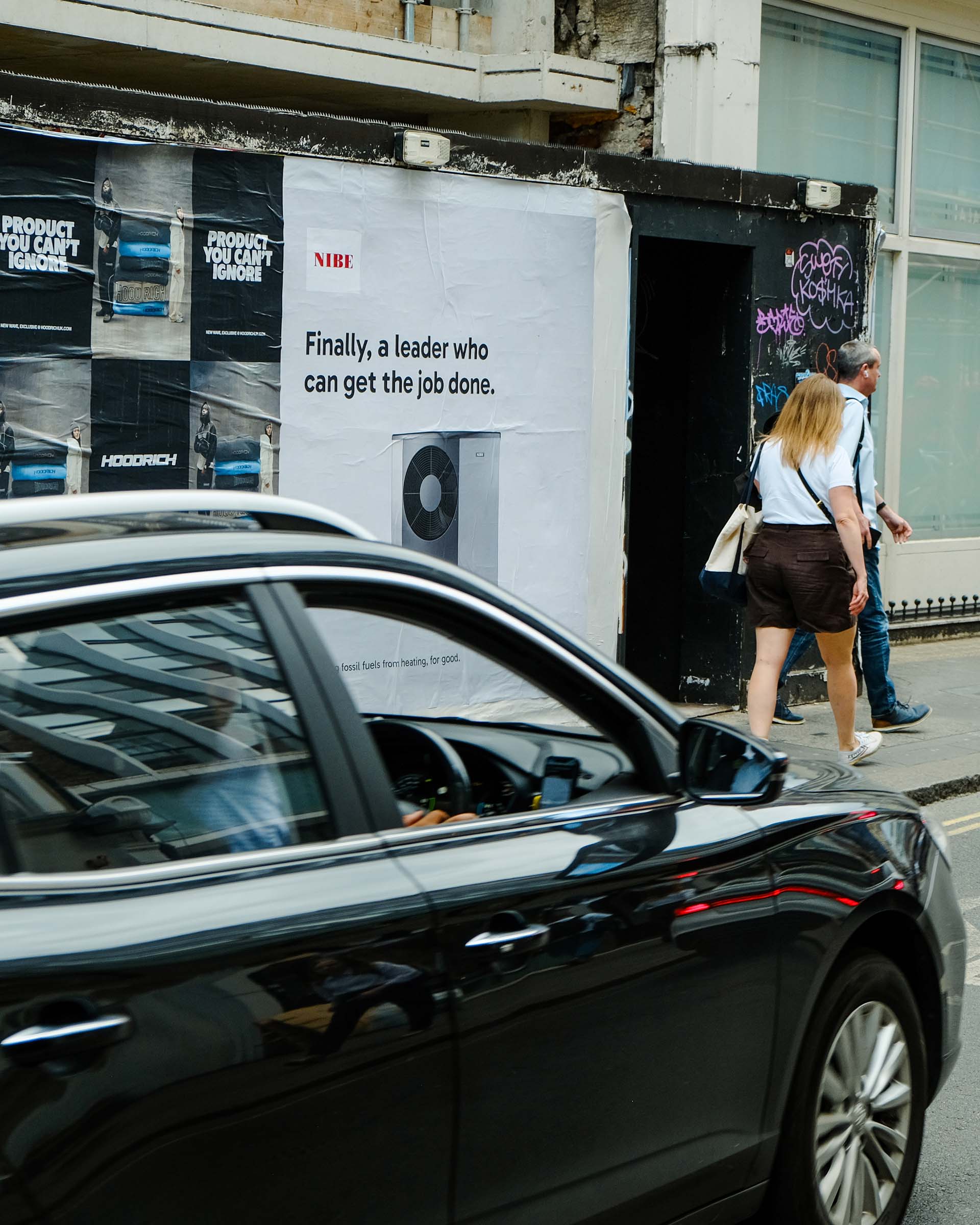Two pedestrians walk past a street wall with a NIBE ad stating, ‘Finally, a leader who can get job done.’