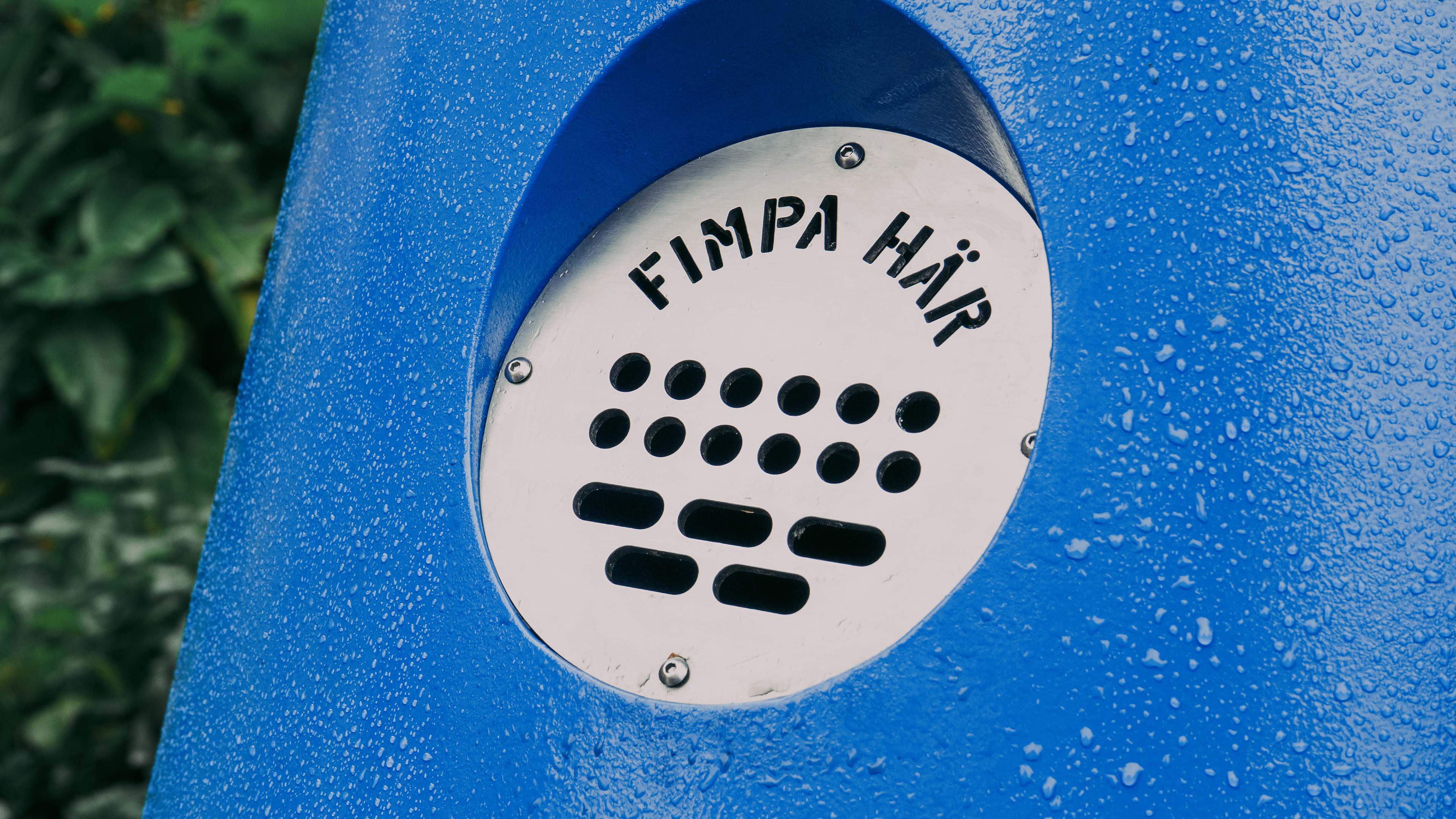 Metal ashtray with the text 'Fimpa här' on a blue surface covered with water droplets.