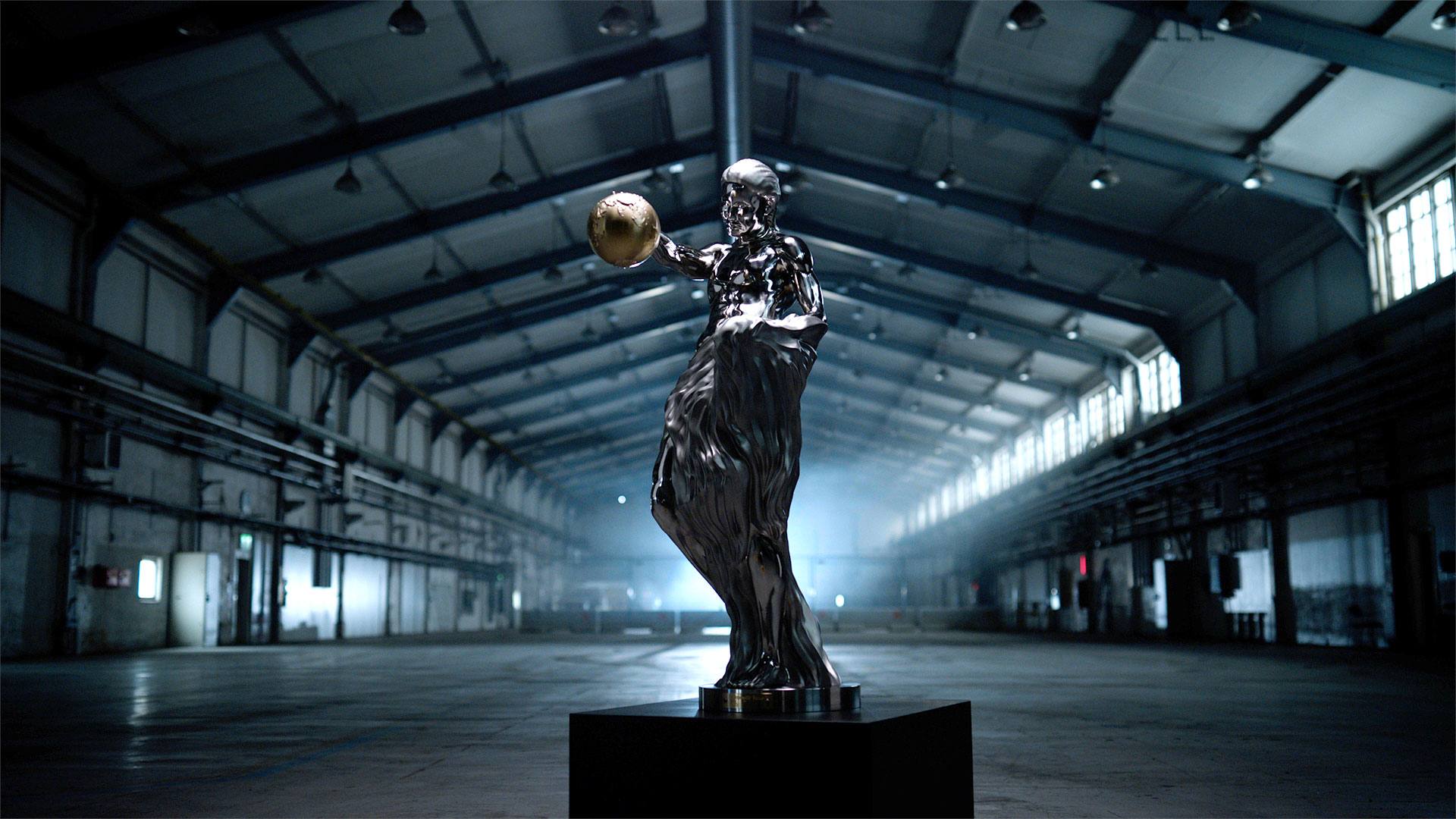 Shiny metallic statue of a figure holding a golden sphere, placed on a pedestal in a large, empty industrial warehouse.
