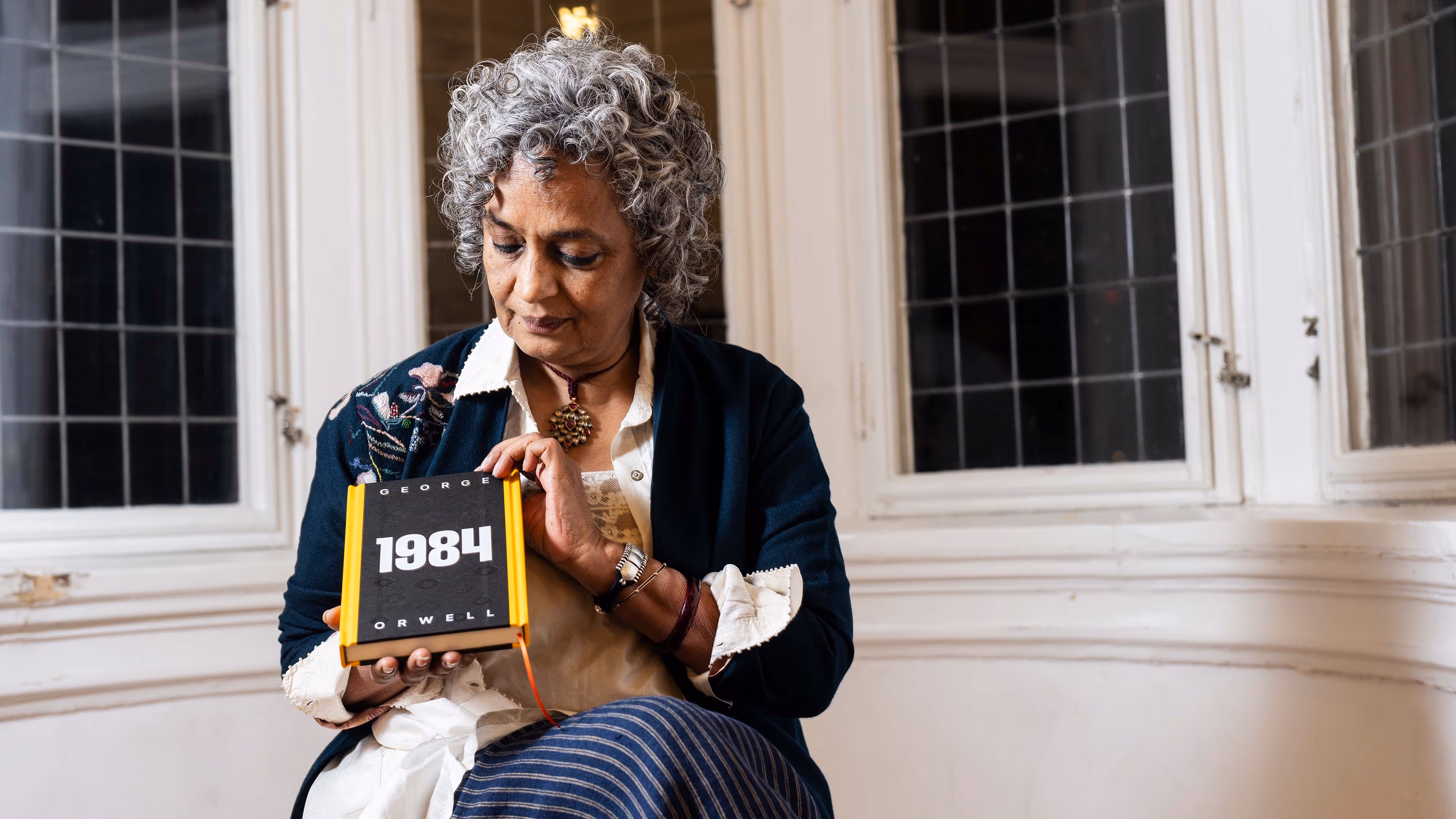Elderly woman with curly gray hair sitting and holding a copy of the book '1984' by George Orwell.