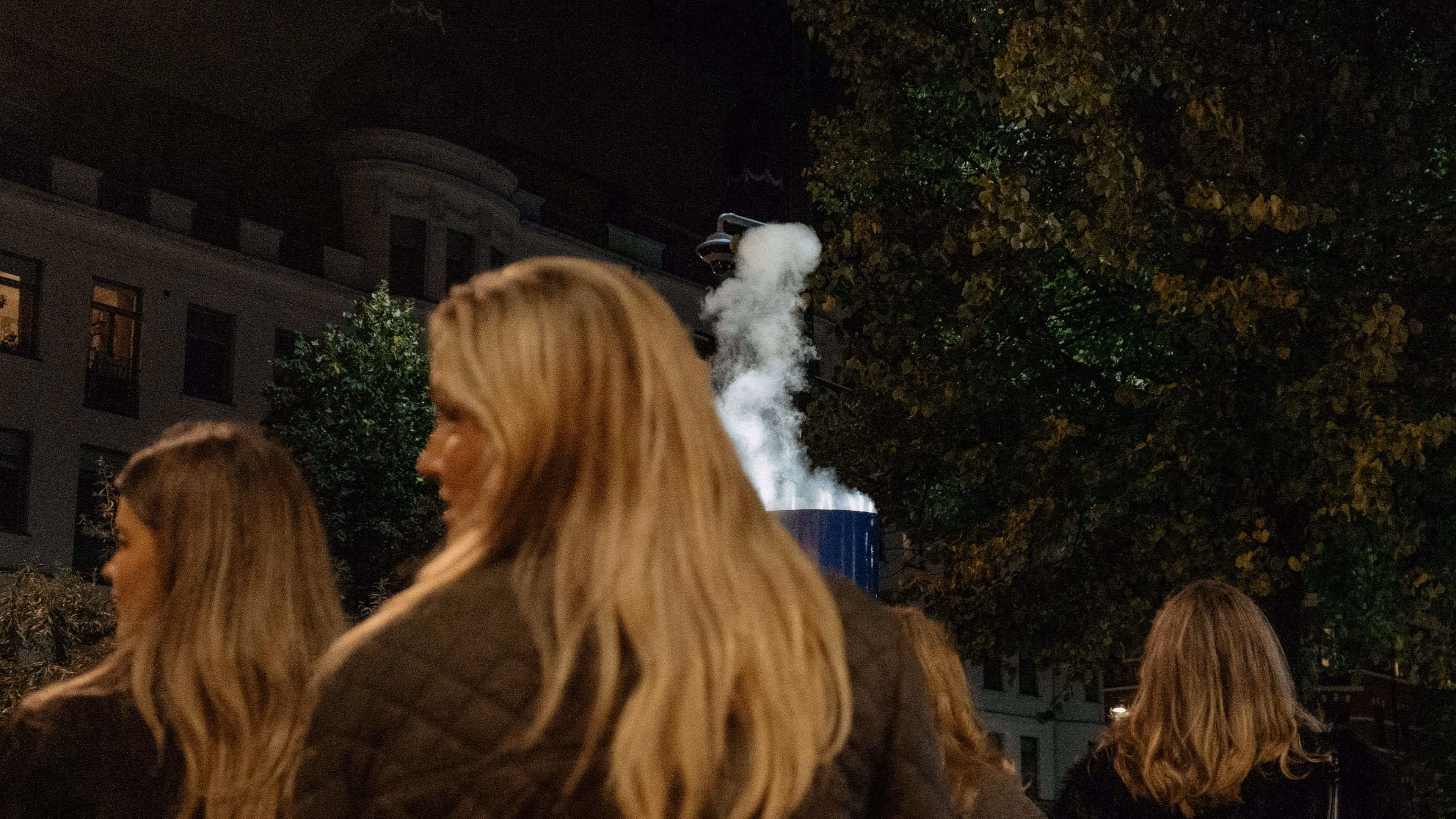 Group of people with long hair walking at night near trees and a building with steam rising from a vent.