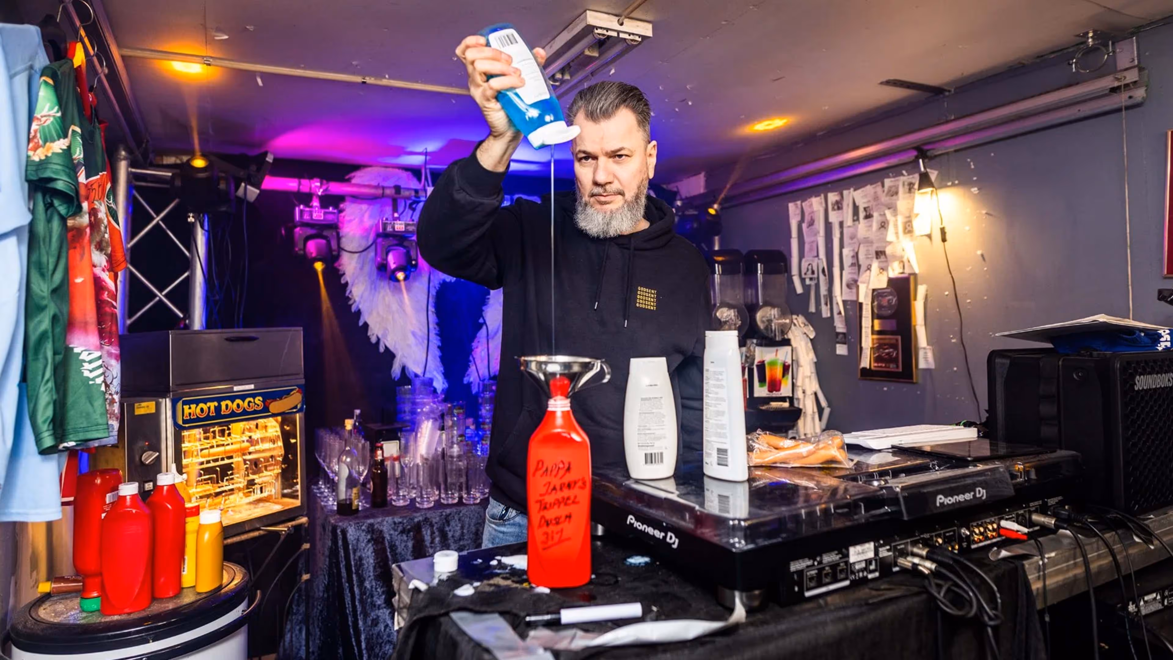 Man with gray beard pouring a liquid from a blue bottle into a red funnel labeled 'Pappa Jarnys Trippel Dusch 317' on a table with DJ equipment and condiments.